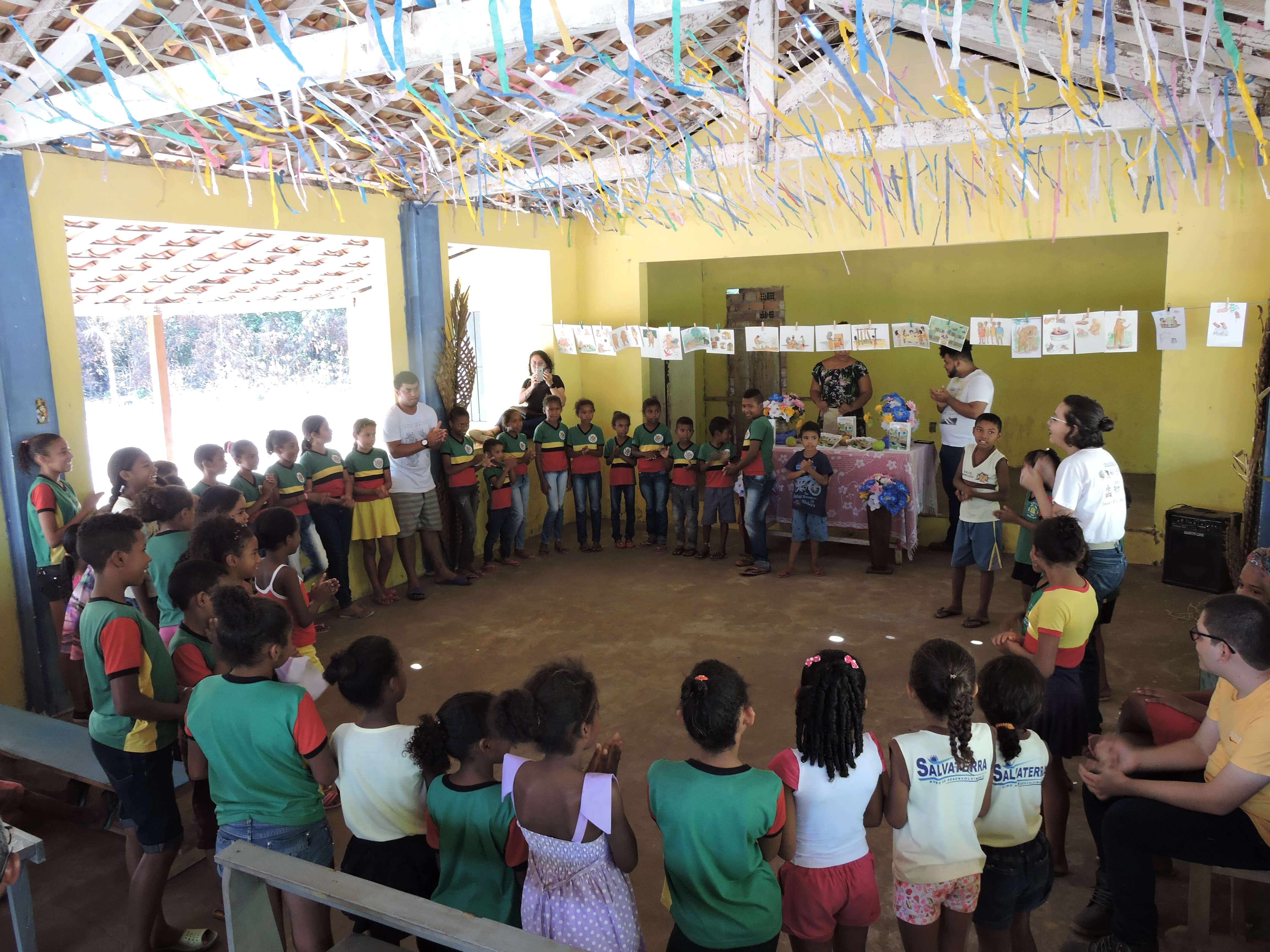 Picture of Beta and their fellow researchers with the quilombola children of both communities studied, they are inside a church that also works as a community center in one of the quilombos and they are also playing ciranda - a game that involves singing and dancing folk/regional songs, in which players are side by side forming a circle - as a way of introducing themselves and also getting to know a little about the childrens perspectives on the activity itself, which was an event to return to the communities part of the research results in the format of a childrens book to be used in the quilombola schools. The activity was set for the morning at the time, we can notice the daylight by looking at the pics.